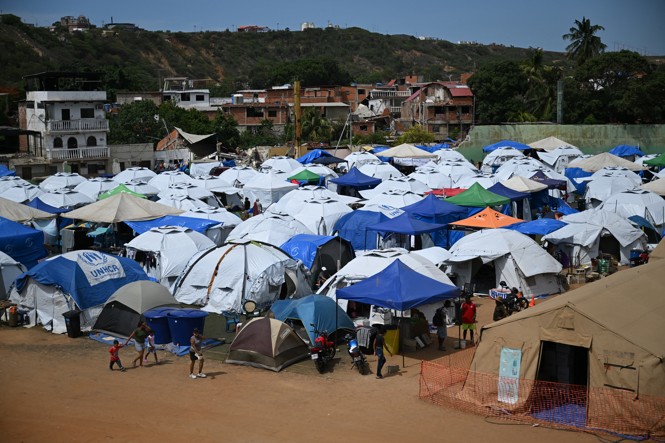 People walking near many tents erected next to a neighborhood of partially collapsed buildings