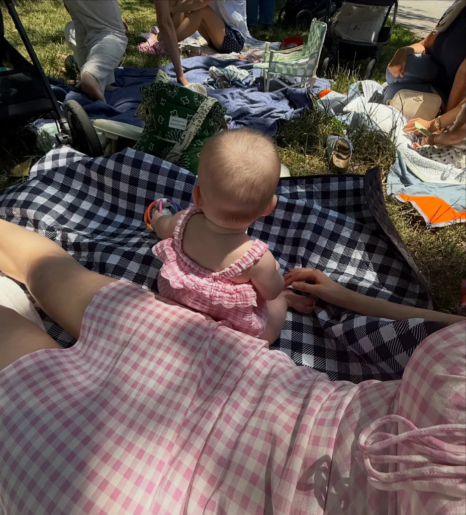A baby in a pink gingham romper sitting on a black and white gingham blanket, surrounded by people picnicking on the grass.