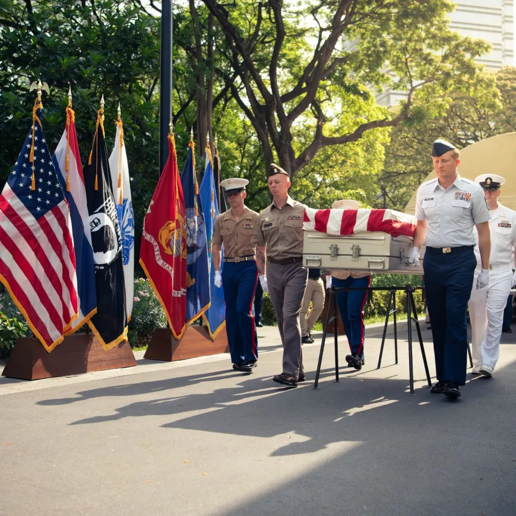 Military personnel carrying a flag-draped casket past a line of flags.