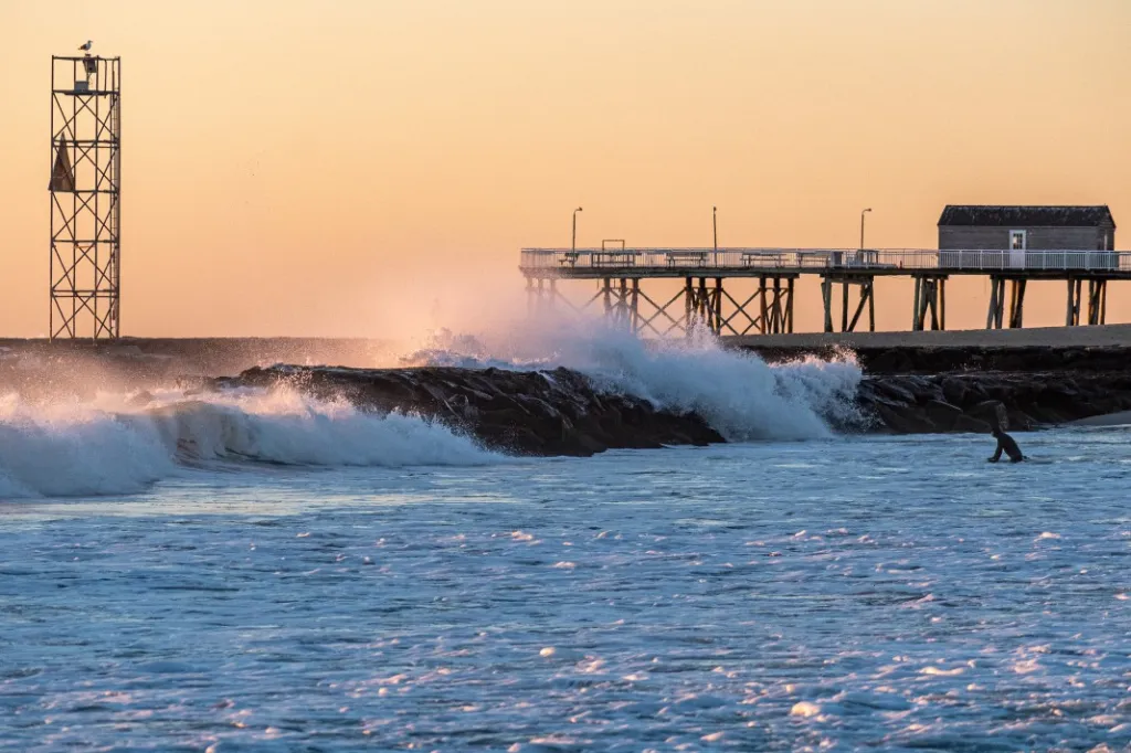 Waves breaking over a rocky jetty at sunrise with a fishing pier in the background.