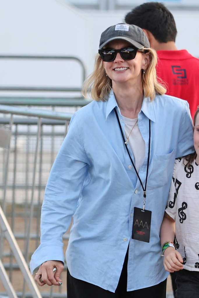 Carey Mulligan smiles while wearing a light blue button-down shirt, a white t-shirt, black sunglasses, and a gray baseball cap with an "AAA" pass hanging around her neck.