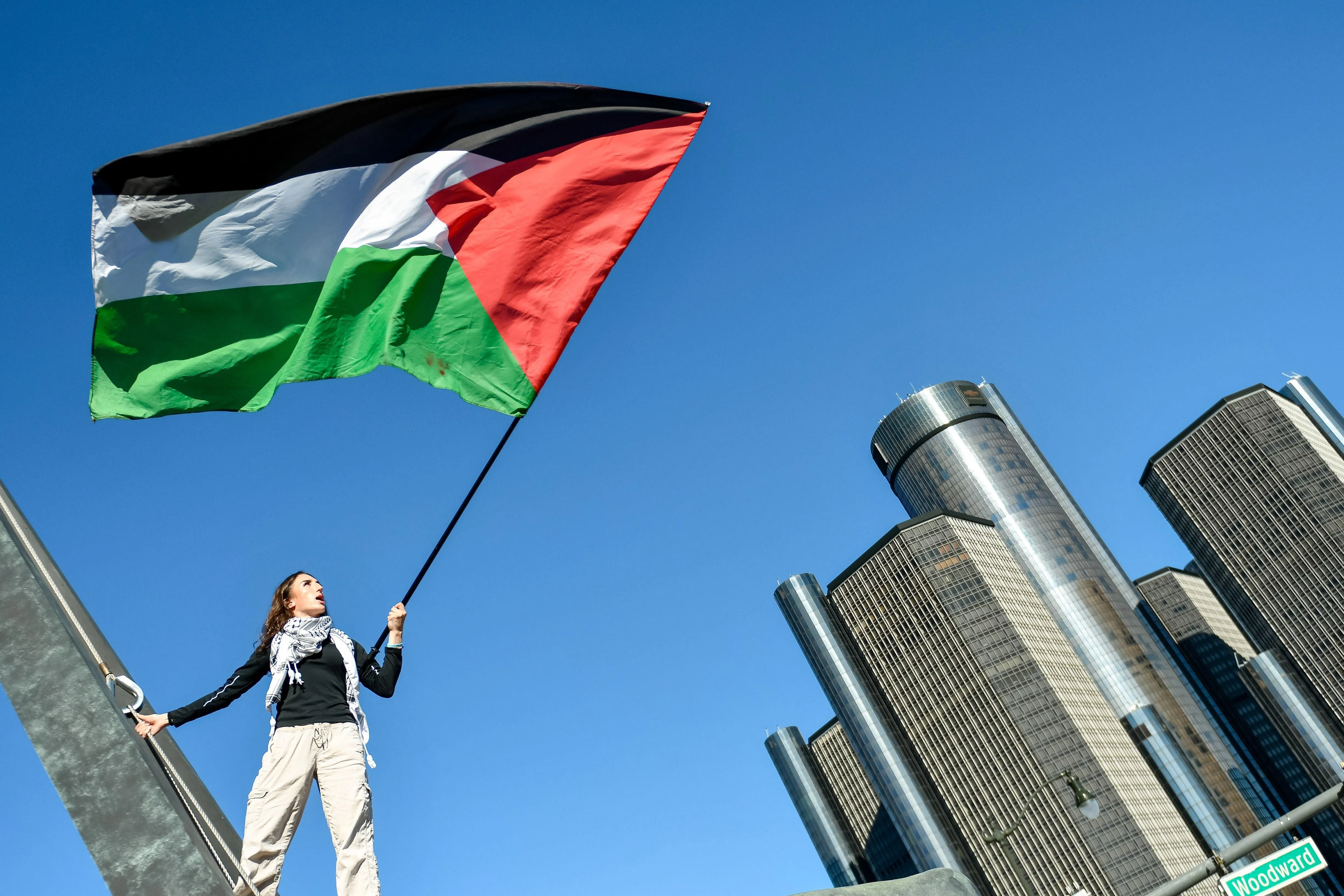 A woman holds a large Palestinian flag in a shot that contrasts the flag with the skyline of downtown Detroit.