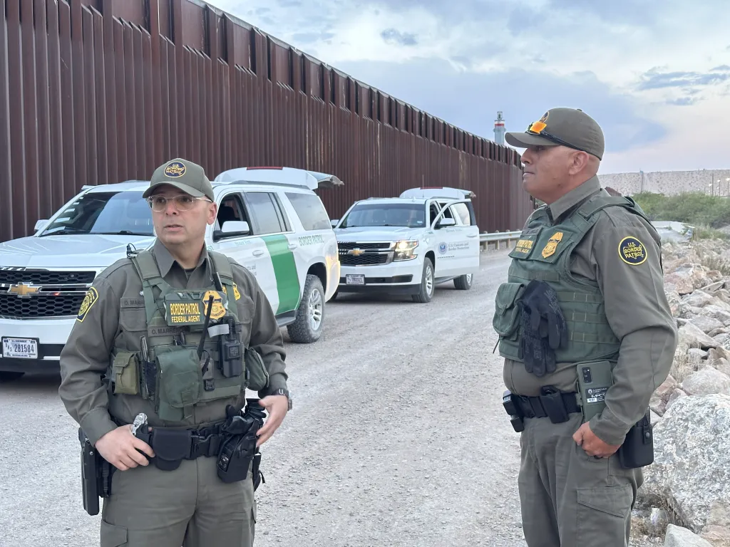 Two Border Patrol agents stand with patrol vehicles near the US-Mexico border wall.