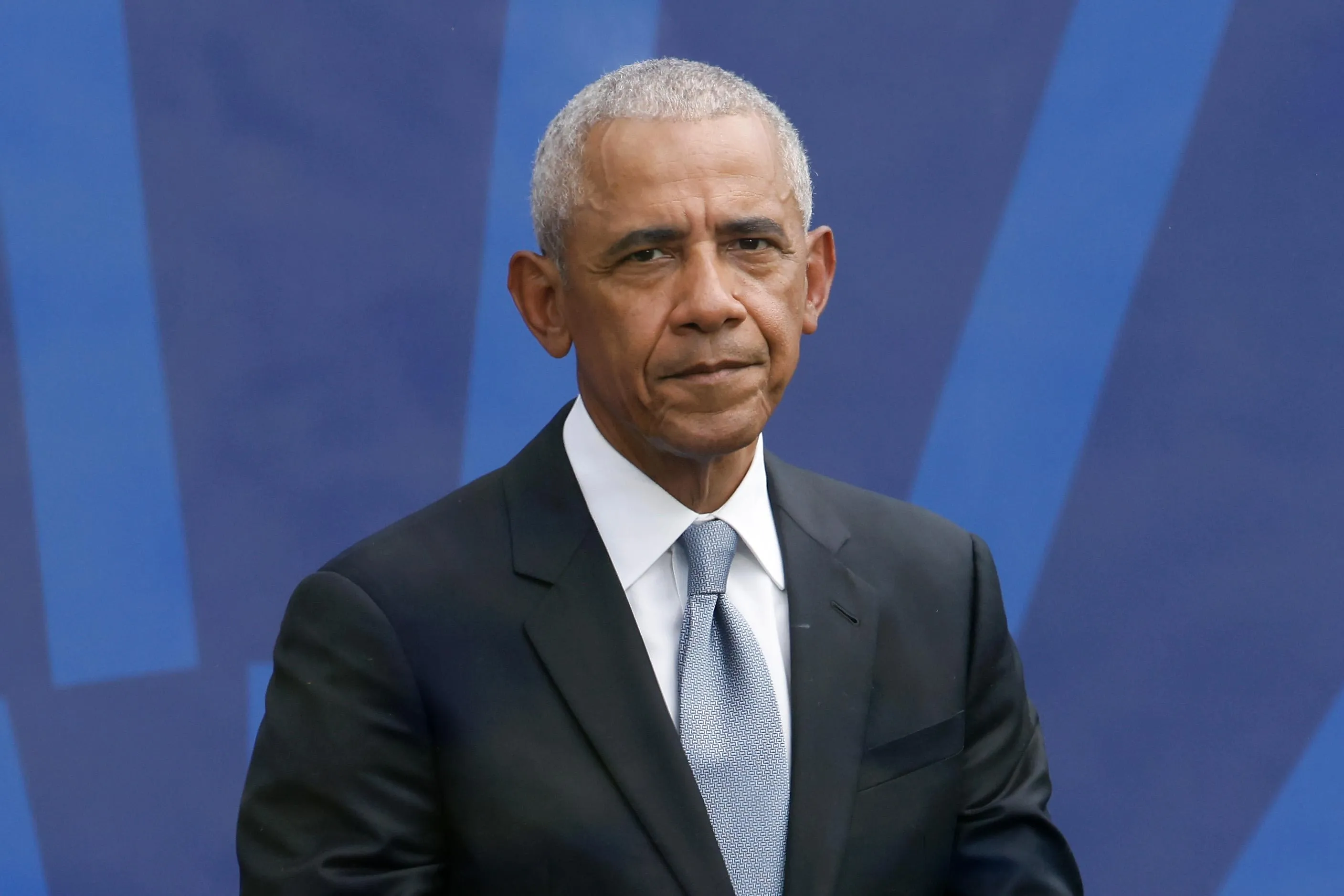 Former President Barack Obama wearing a black suit, light blue tie, and sitting against a blue background.