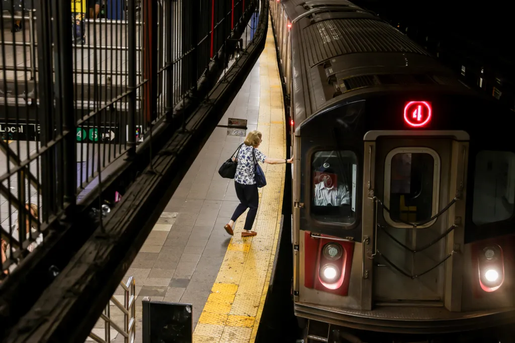 A woman in a floral shirt and dark pants stands on a subway platform next to a train with a red "4" sign.