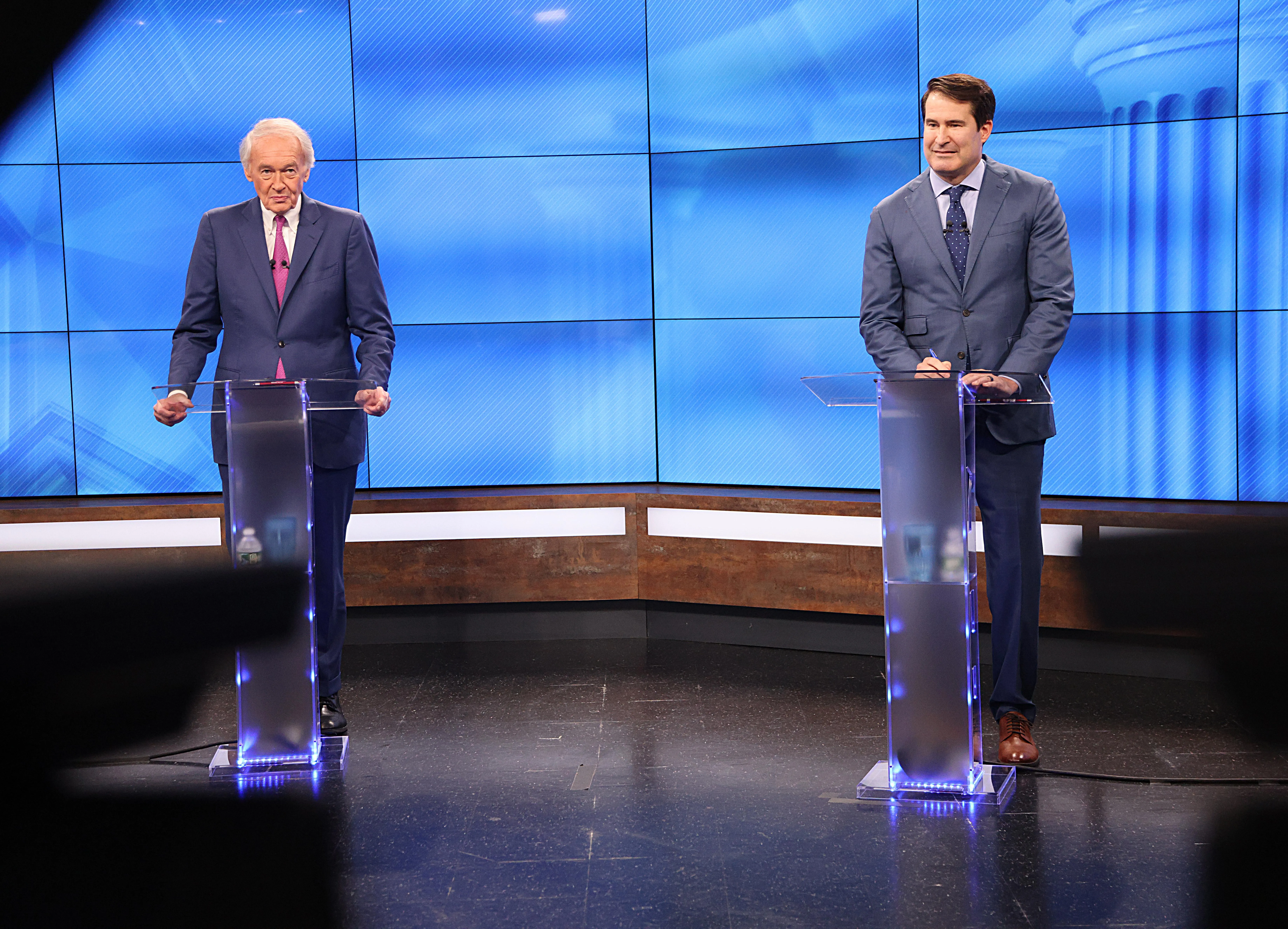 two men stand behind purple-lighted podiums and in front of a blue screen