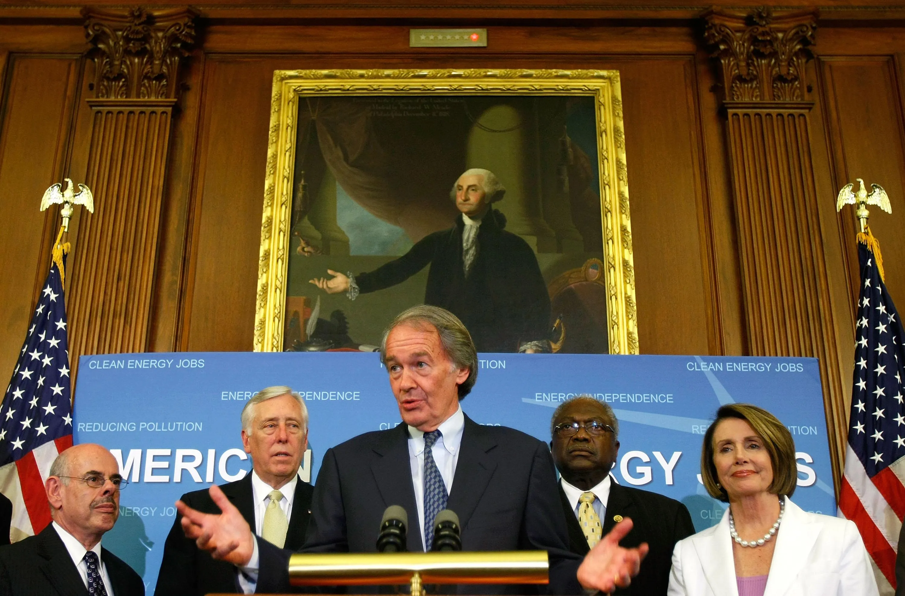 a man stands behind a podium; in the background stand three other men and a woman; they are standing in front of a blue background with American flags on either side