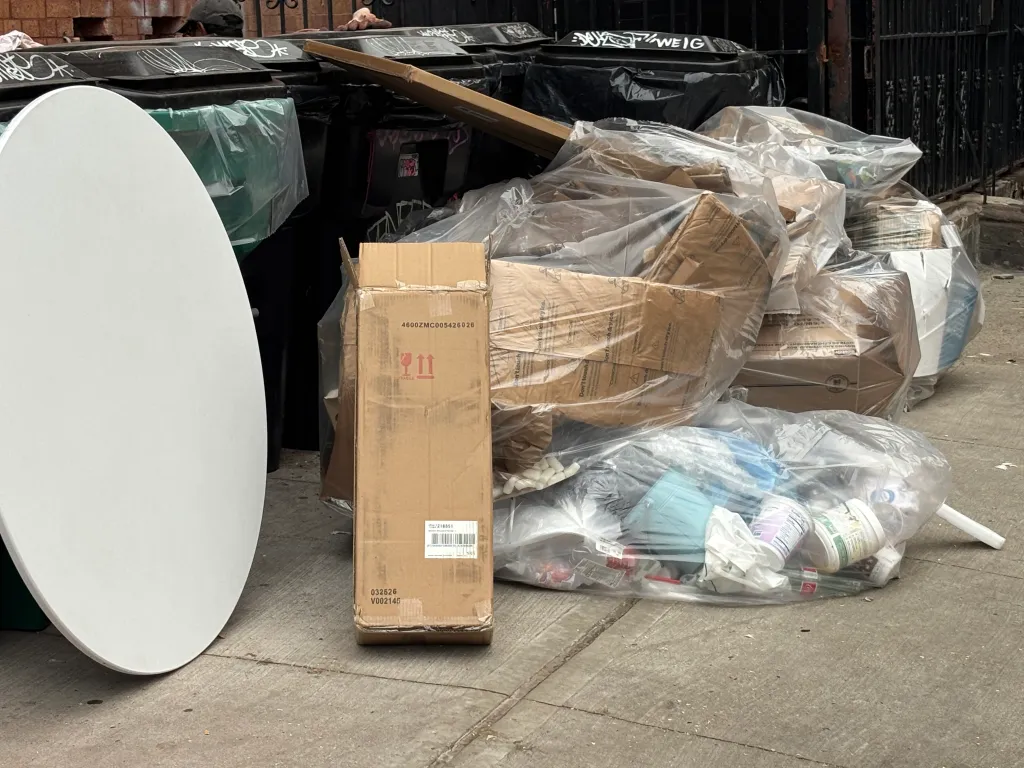 A tall, thin cardboard box with "FRAGILE" written on it stands next to a large pile of clear plastic bags filled with boxes and trash.