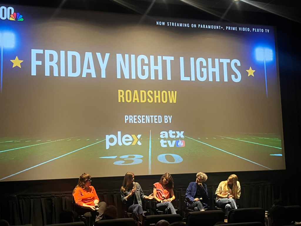 A screen displaying "Friday Night Lights Roadshow" and five panelists speaking on stage.