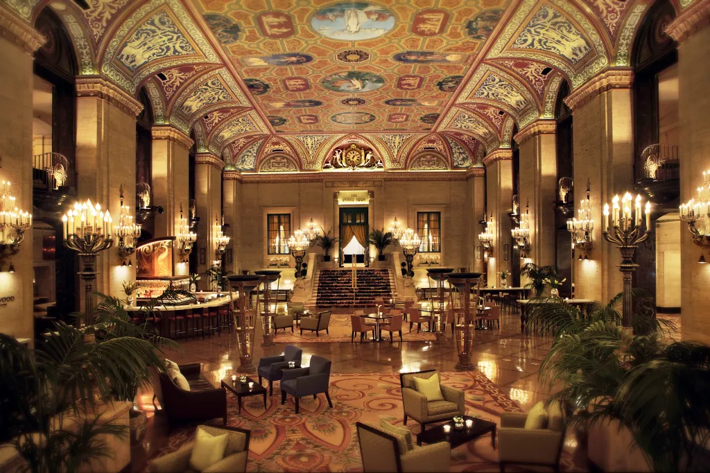 The luxurious Huber Lobby of the Palmer House Hilton, with a grand painted ceiling, elegant chandeliers, a bar area, and seating arranged on a patterned rug.