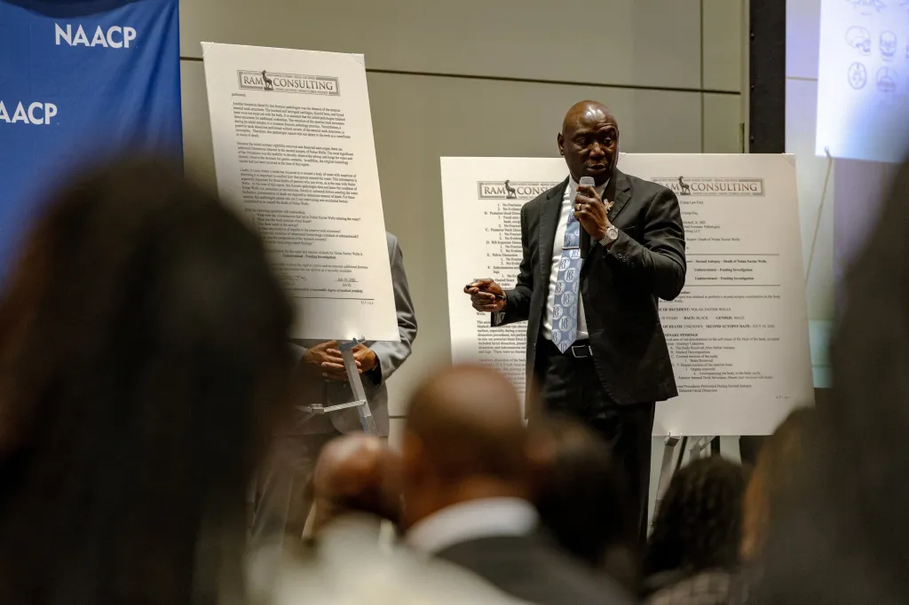 A lawyer speaks into a microphone in front of two display boards detailing autopsy results in the Nolan Wells case.