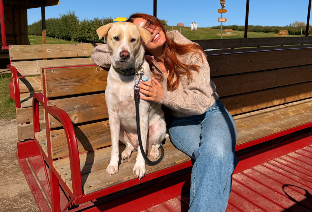 A young woman in a beige sweater and jeans smiles while hugging her dog sitting on a wooden bench.