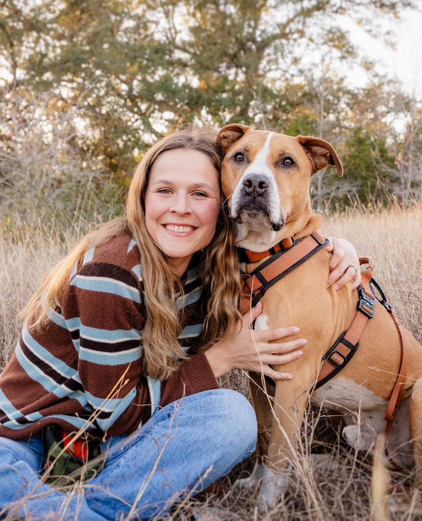 A woman in a striped sweater and jeans smiles while hugging a brown and white dog in a field of dry grass.