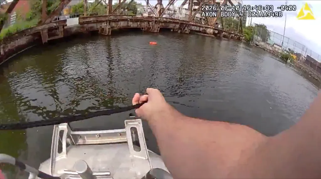 A person's hand pulling a rope from a boat in the water, with a life ring floating in the distance near a bridge.