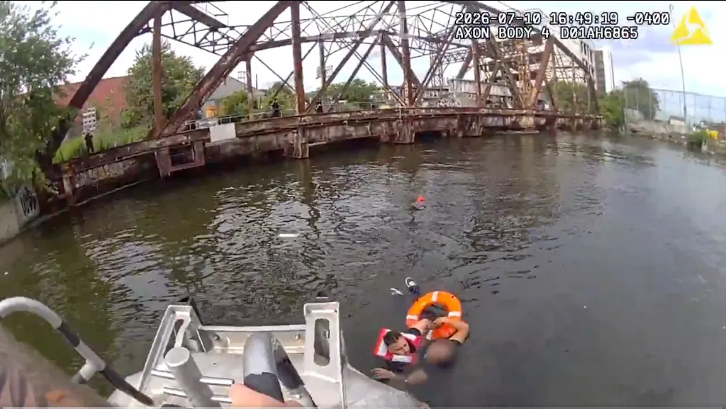 Two people in life vests in the water next to a boat, with a large metal bridge in the background.
