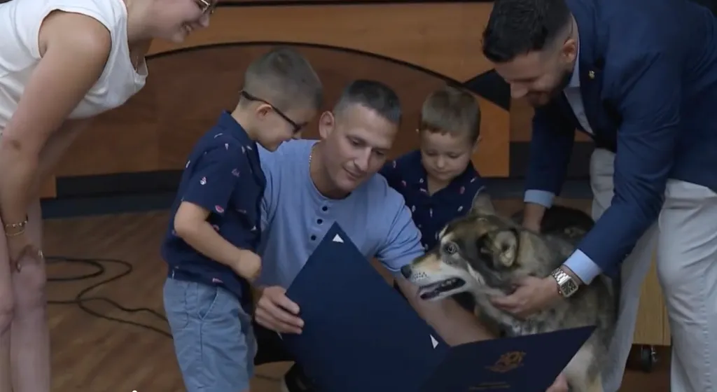 A man sitting on the floor with two boys looking at a blue folder, while another man pets a dog beside them.