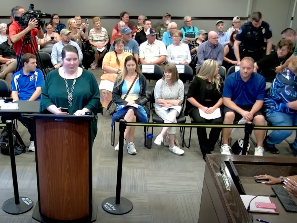 A woman in a green shirt and long necklace speaks at a podium in a public meeting with an audience seated behind her.