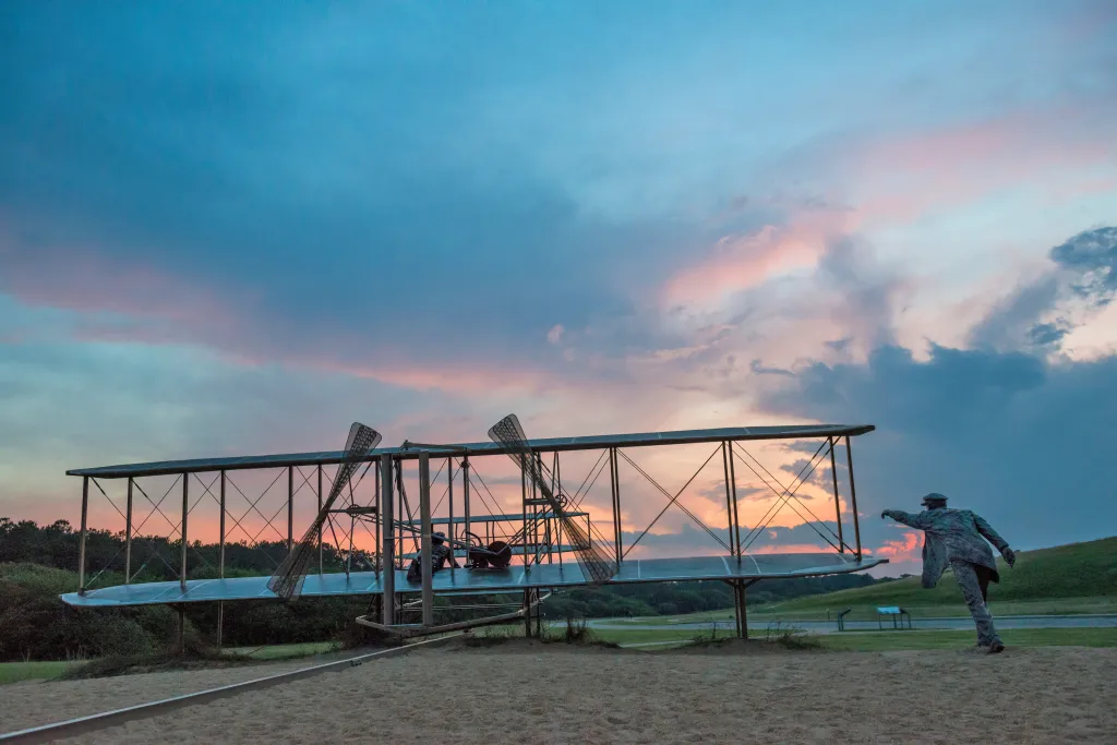 The Wright Flyer replica and a statue of a person on the sand at the Wright Brothers National Memorial in Kill Devil Hills, North Carolina, at sunset.