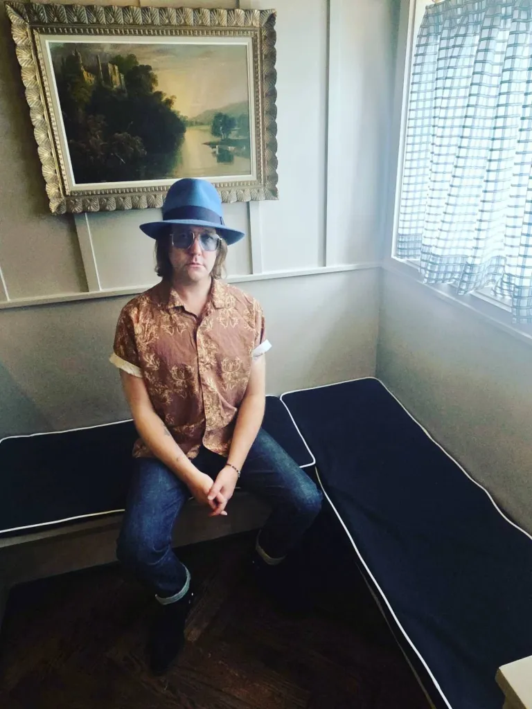 Aaron Lee Tasjan sits wearing a blue fedora, sunglasses, patterned shirt, and jeans.