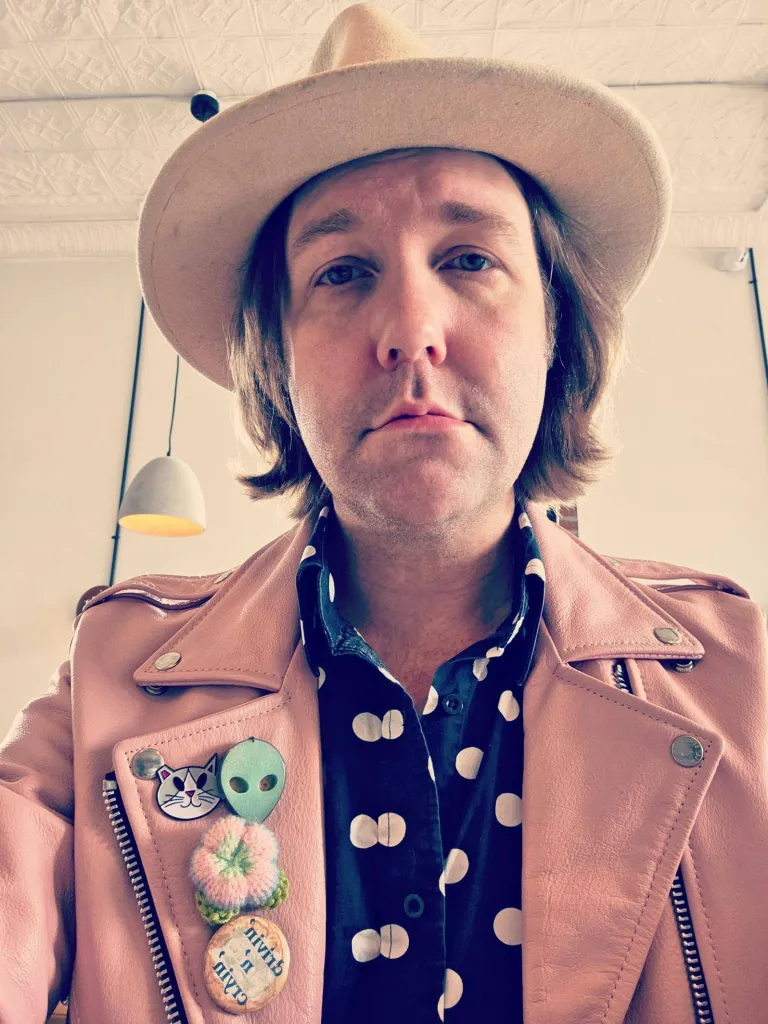Aaron Lee Tasjan looking at the camera wearing a hat, a polka dot shirt, and a pink leather jacket with pins.