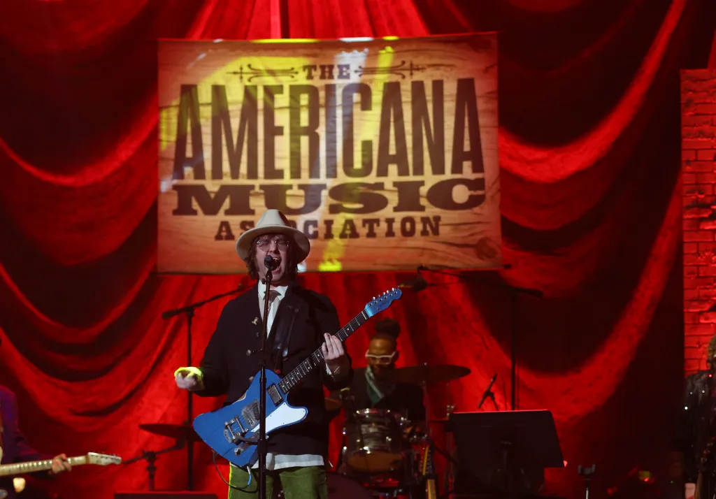 Aaron Lee Tasjan performs on stage, wearing a fedora and glasses, playing a blue electric guitar in front of a red curtain and "The Americana Music Association" banner.