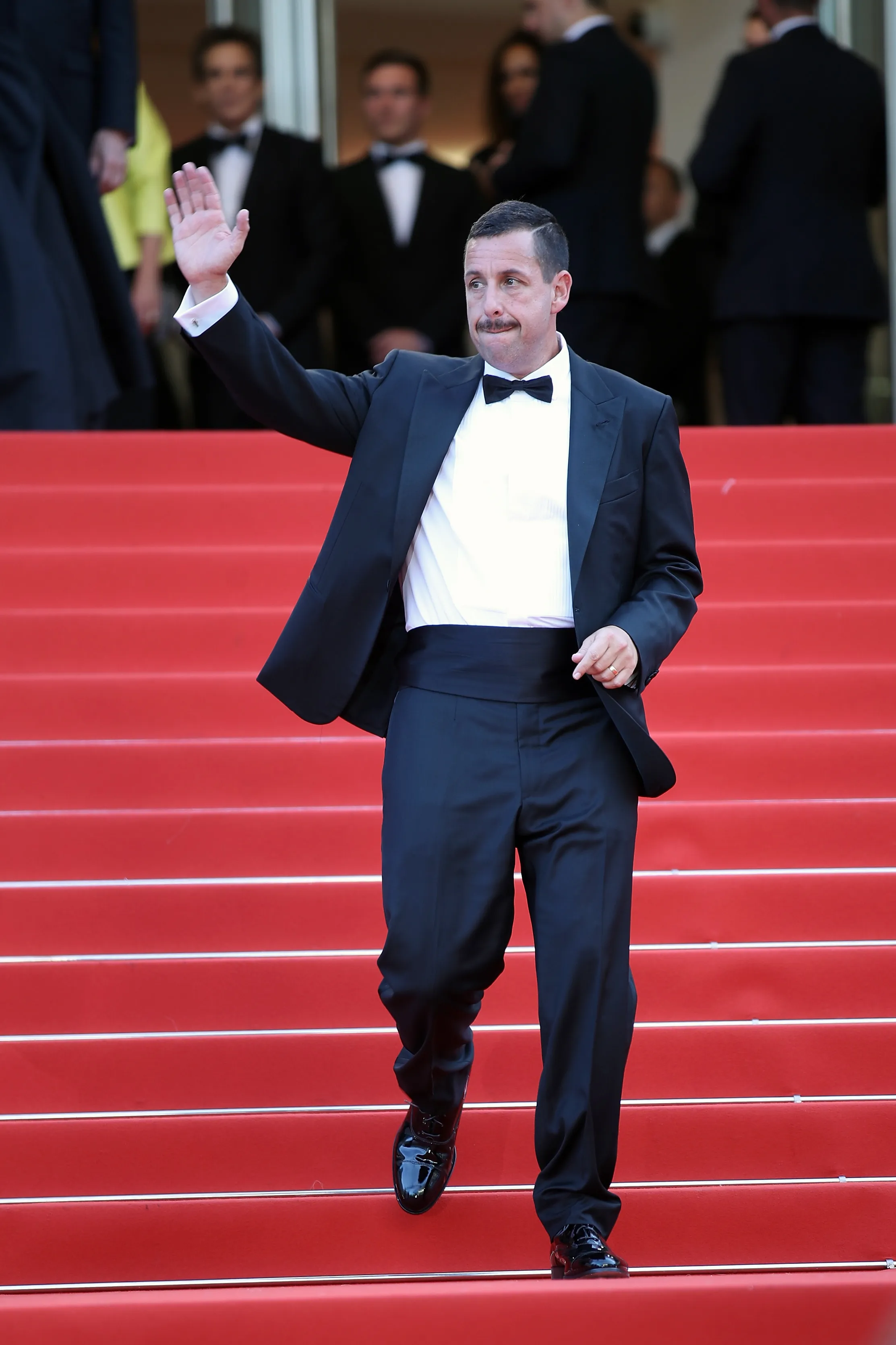 Adam Sandler on a red carpet, wearing a tuxedo and waving.