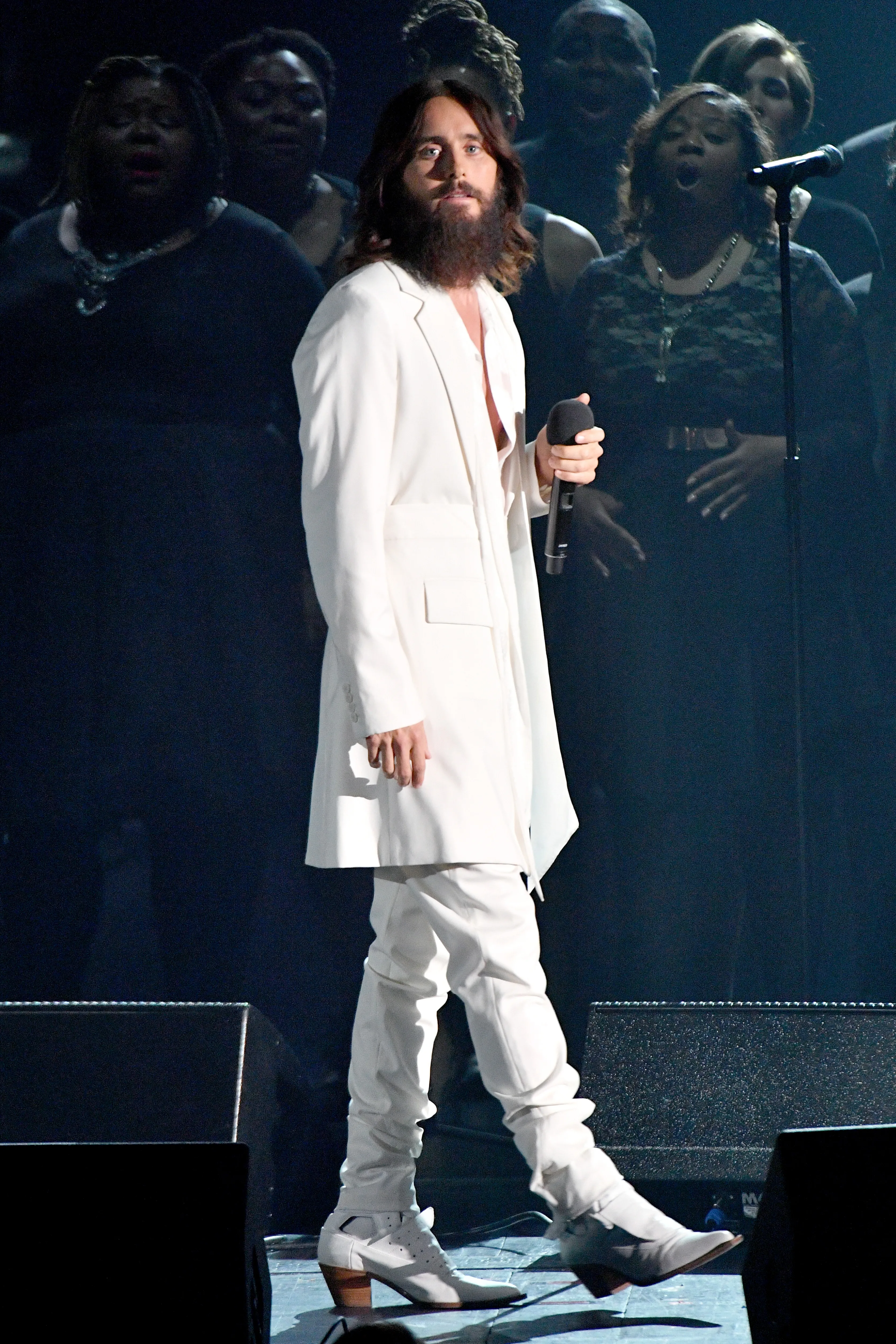 Jared Leto performing onstage in a white suit with a choir in the background.