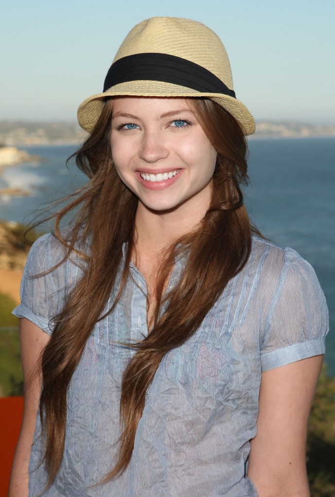 Actress Daveigh Chase smiles, wearing a straw fedora and a pinstriped shirt, with the ocean in the background.