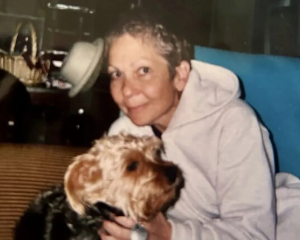 Ada Beth Kaplan Moore in a light-colored hooded sweatshirt, smiling and holding a small dog.