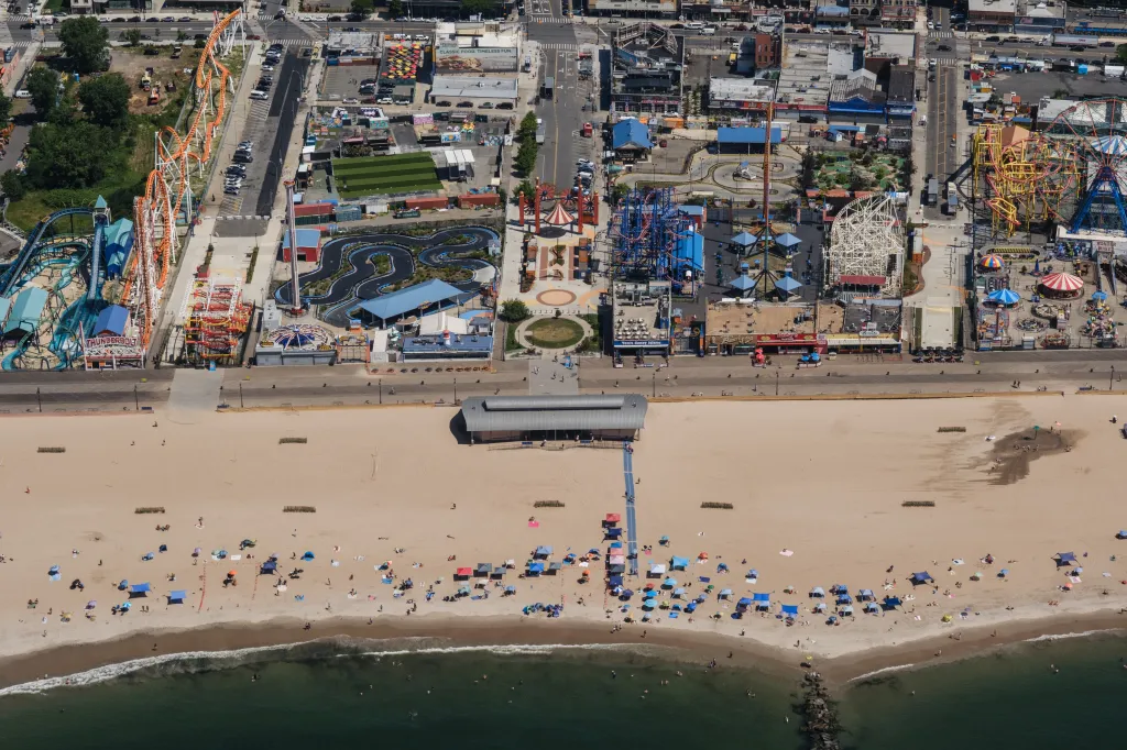 Aerial view of Coney Island, Brooklyn, showing the beach with people, an amusement park with roller coasters and rides, and streets with buildings and cars.