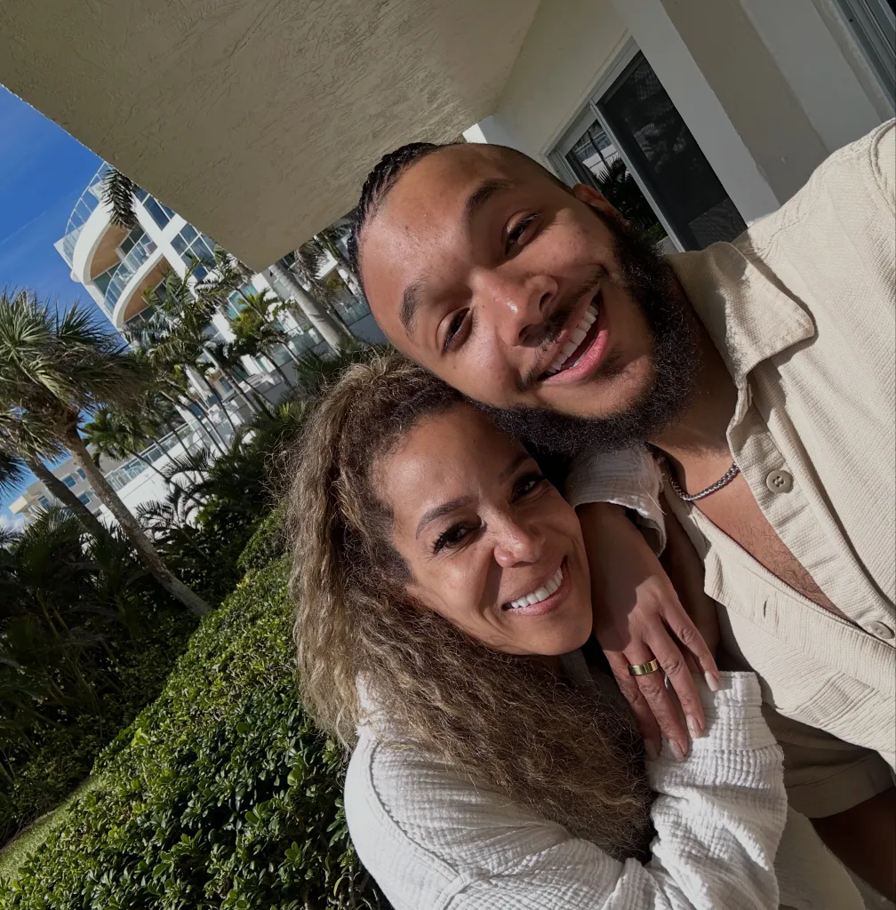 Sunny Hostin and her son Gabriel Hostin (aka Gabe Hostin) smiling in a selfie, with palm trees and a building in the background.