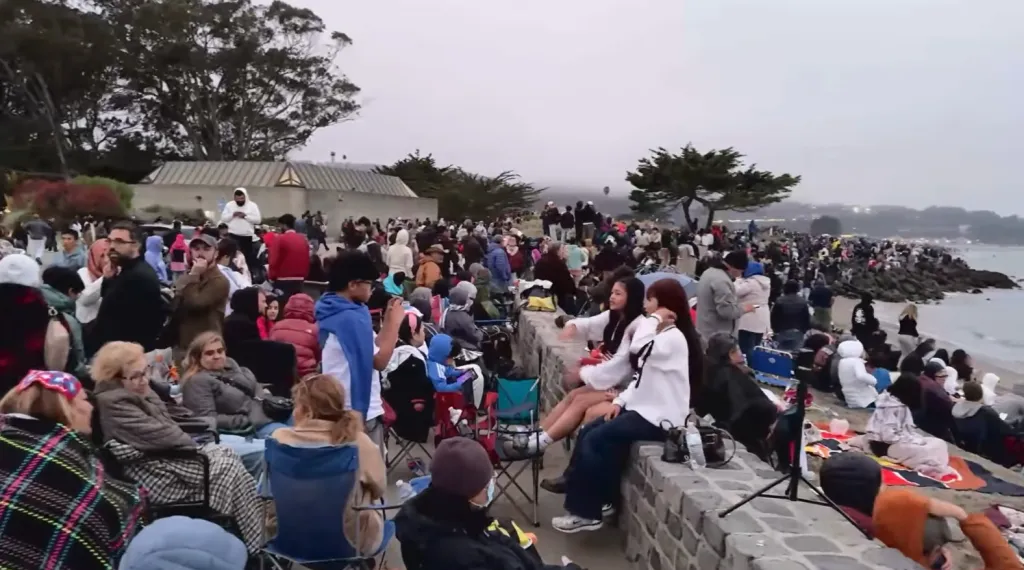 A large crowd of people gathered on a cloudy day, some sitting in chairs or on blankets, facing the ocean.
