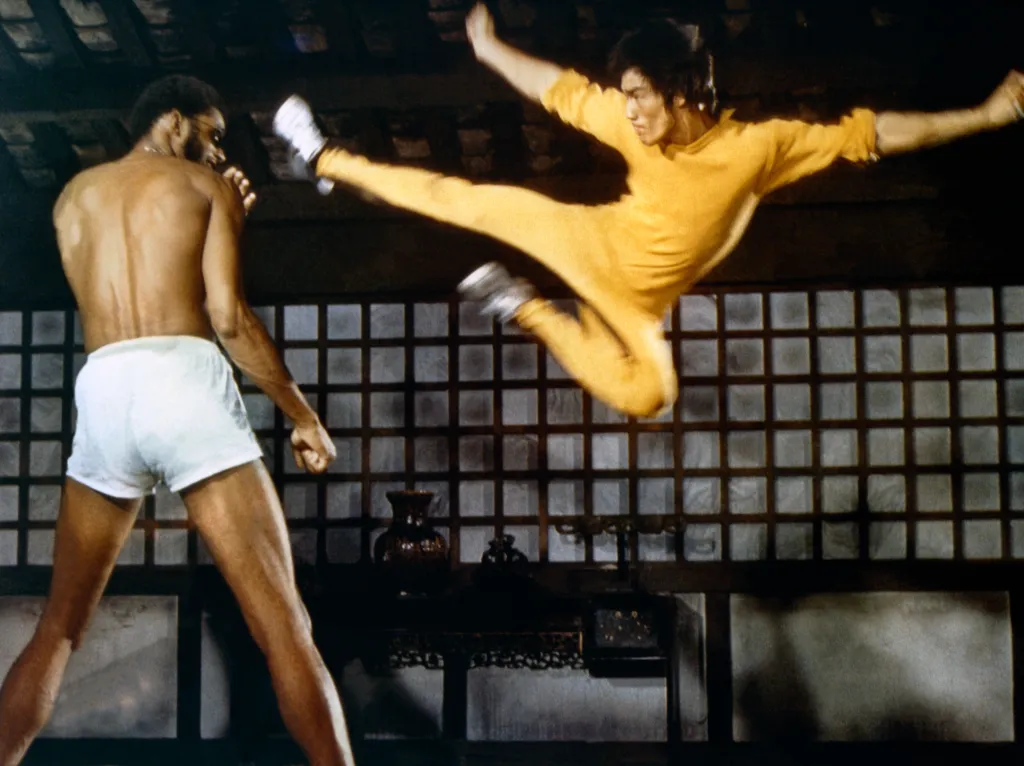 Bruce Lee in a yellow jumpsuit performs a flying kick at Kareem Abdul-Jabbar in white shorts.