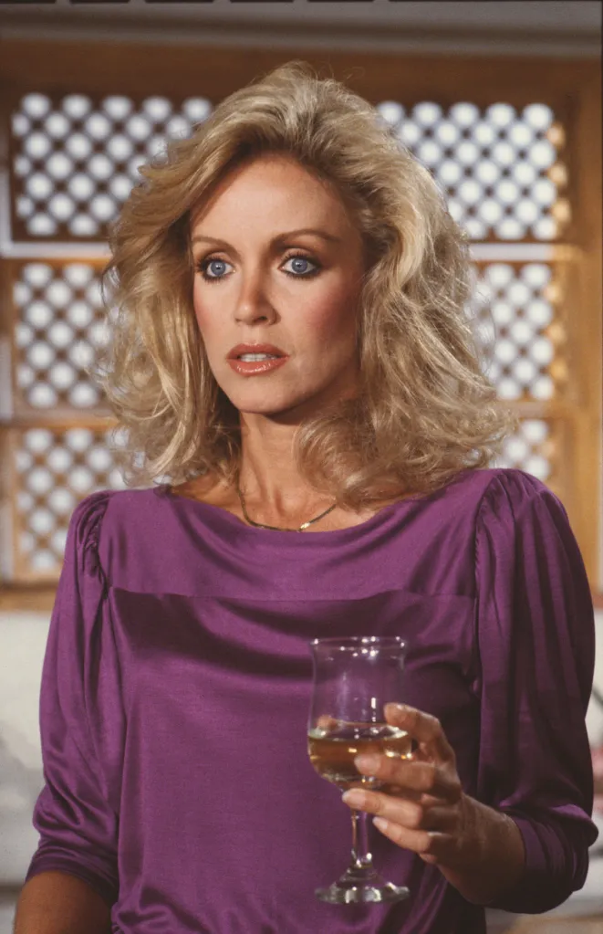 Actress Donna Mills in a purple top holds a glass of wine.