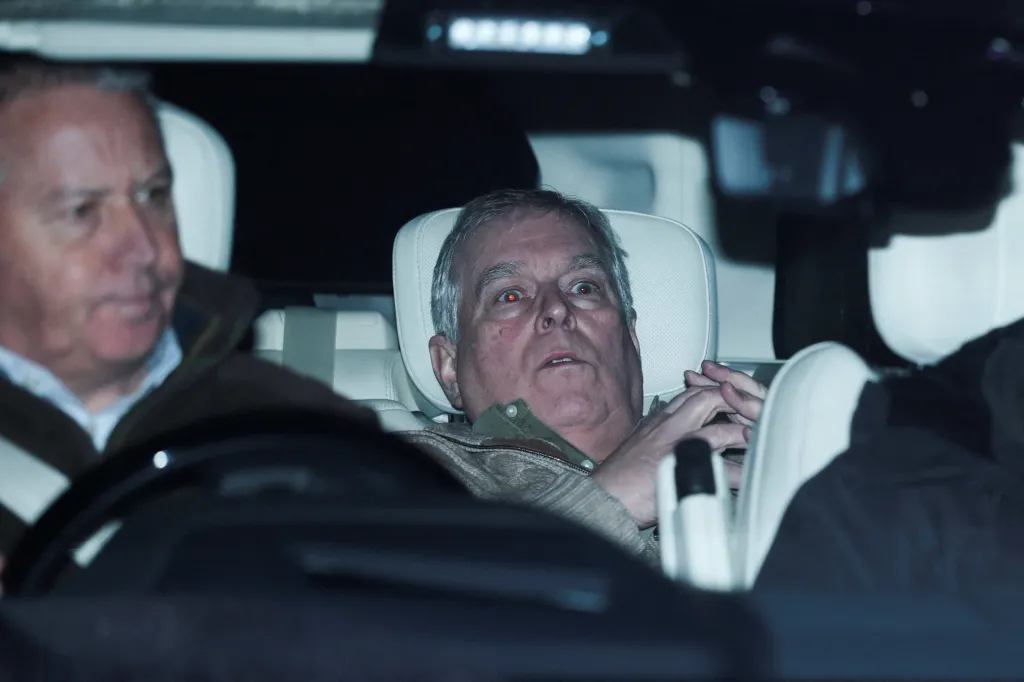 Prince Andrew, dressed in an olive green shirt and patterned jacket, sits in the back of a car with wide eyes and clasped hands.