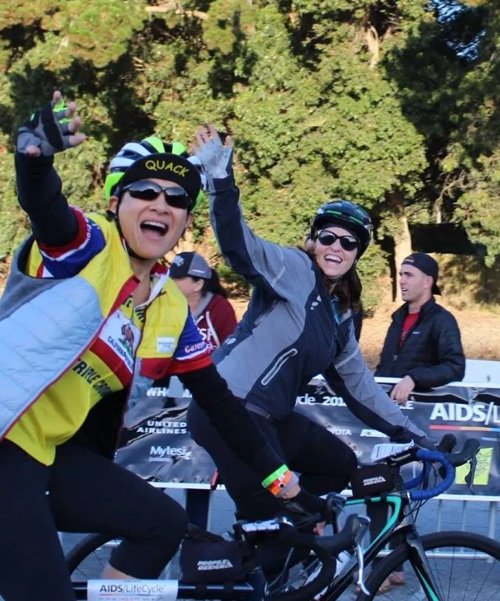 Angela Calvillo and Elaine Forbes, wearing helmets and sunglasses, riding bicycles and waving.