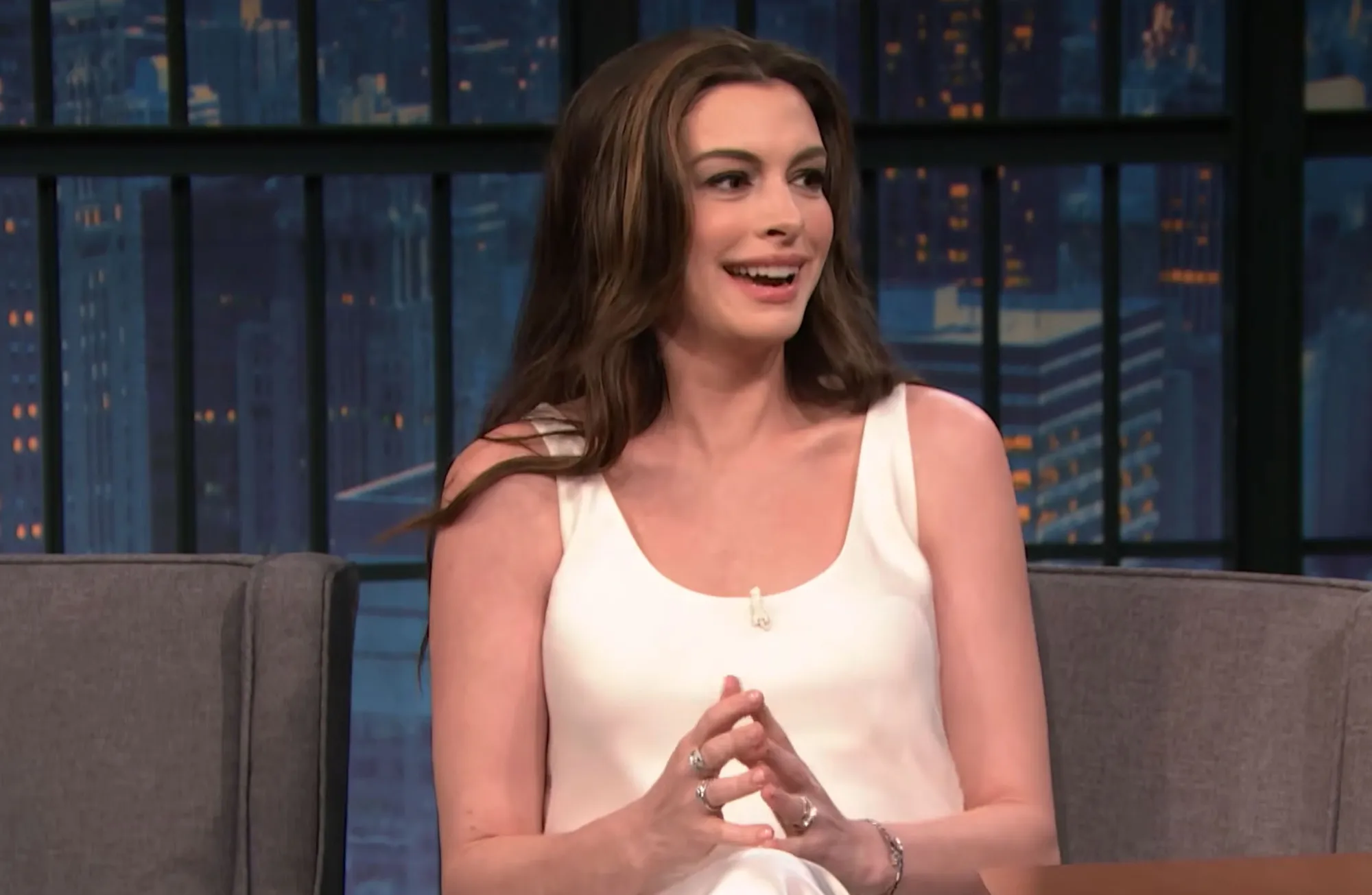 Anne Hathaway seated, smiling, in front of a city background.
