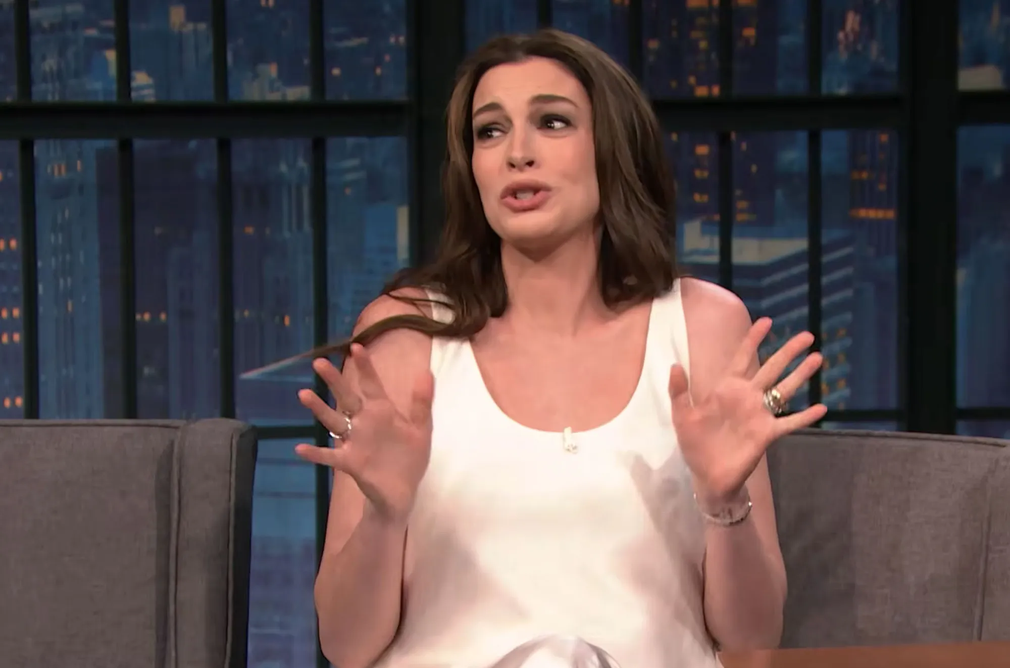 Anne Hathaway in a white tank top speaking on a talk show, gesturing with both hands.