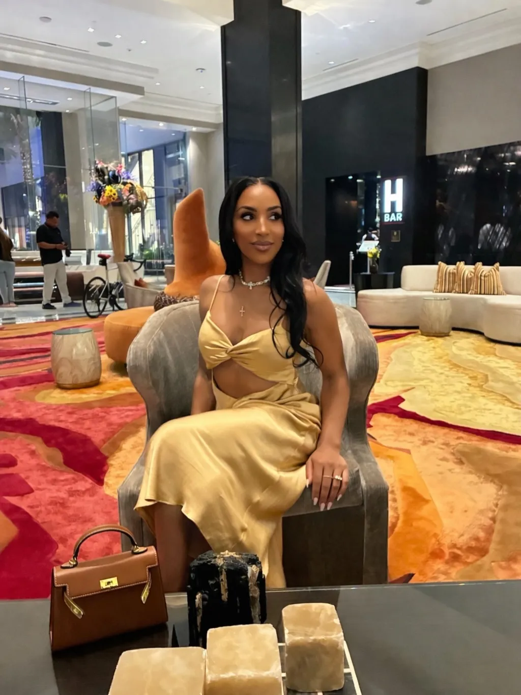 Annemarie Wiley wearing a golden cutout dress, sitting in a chair in a hotel lobby.