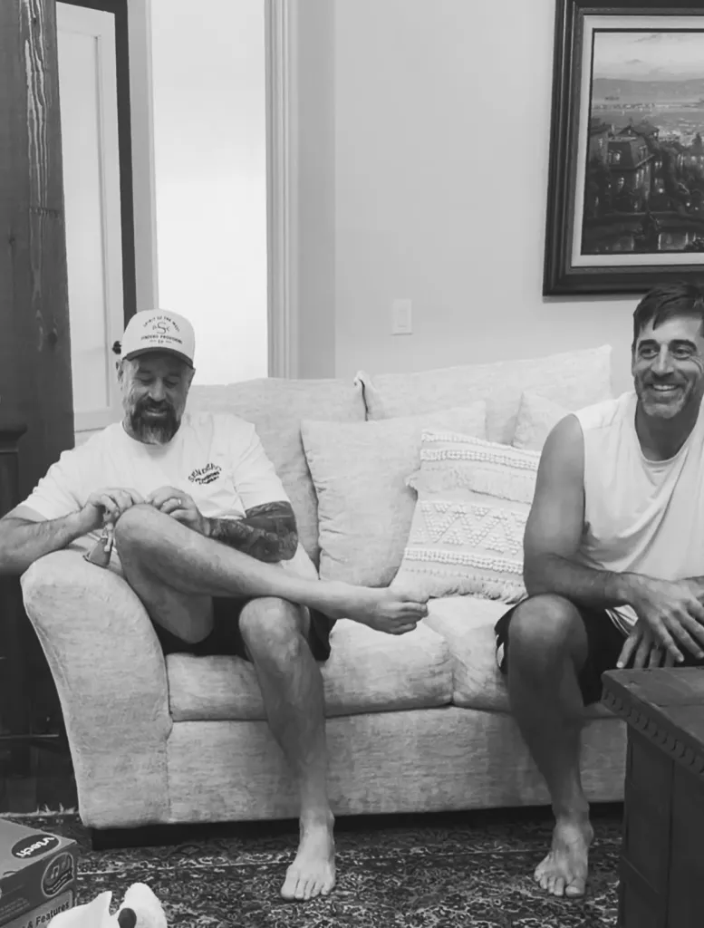 Aaron Rodgers and another man smiling while relaxing on a couch.