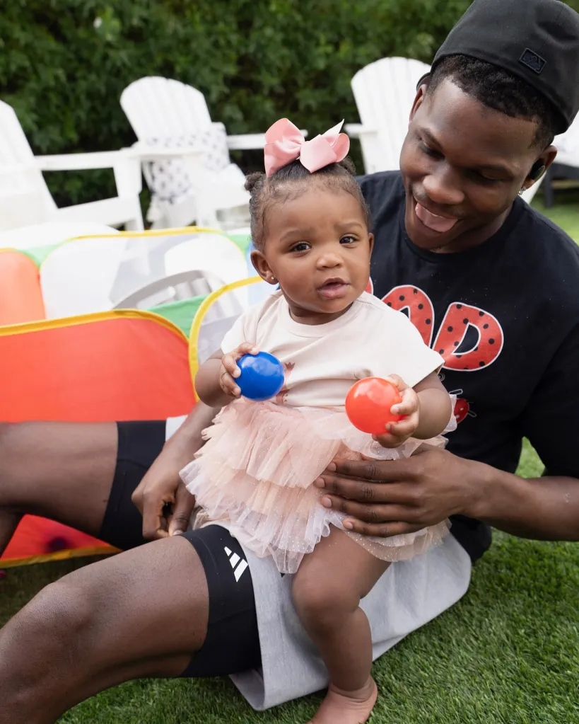 Anthony Edwards holds his daughter Aislynn on his lap as she holds a blue and orange ball.