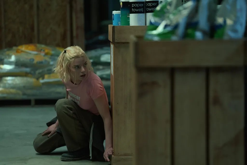 Anya Taylor-Joy crouches behind a wooden barrier, looking over her shoulder with an alarmed expression.