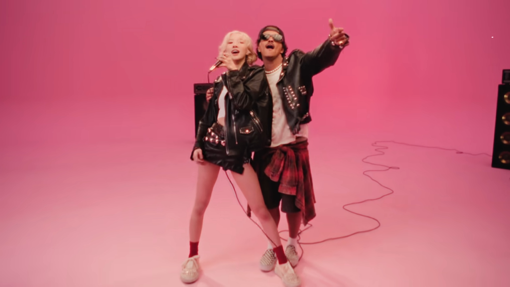 Two singers, a woman and a man, wearing punk rock outfits, perform against a pink background.