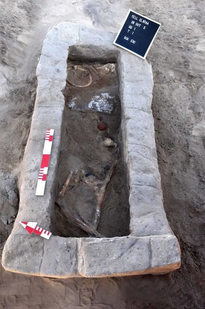 An ancient human skeleton and red pottery fragments partially covered in dirt inside a stone sarcophagus.