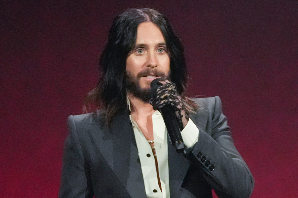 Jared Leto speaking at CinemaCon while wearing a suit, a light shirt, and a black lace glove, holding a microphone.