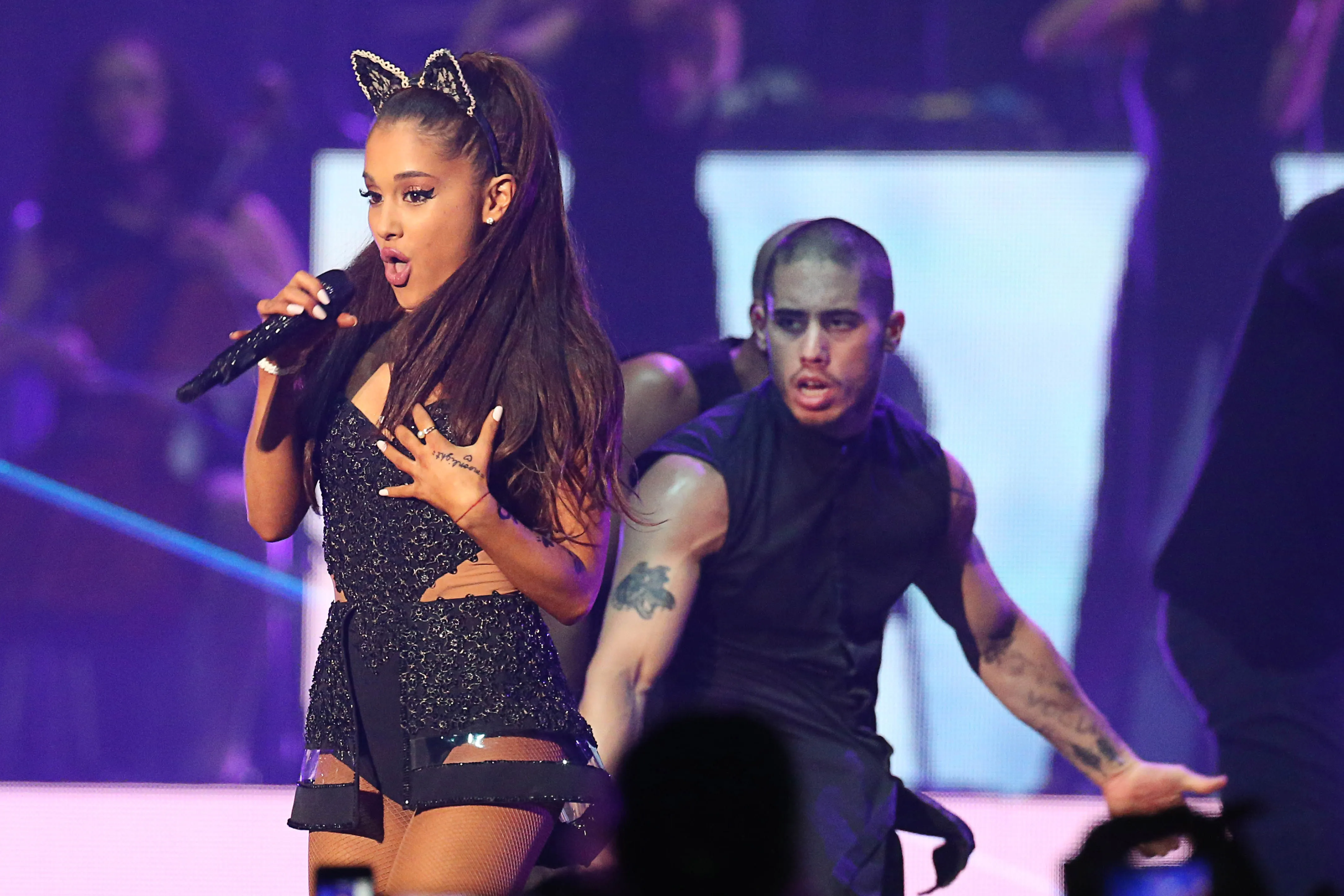 Ariana Grande singing into a microphone on stage, with a dancer and colorful lighting in the background.