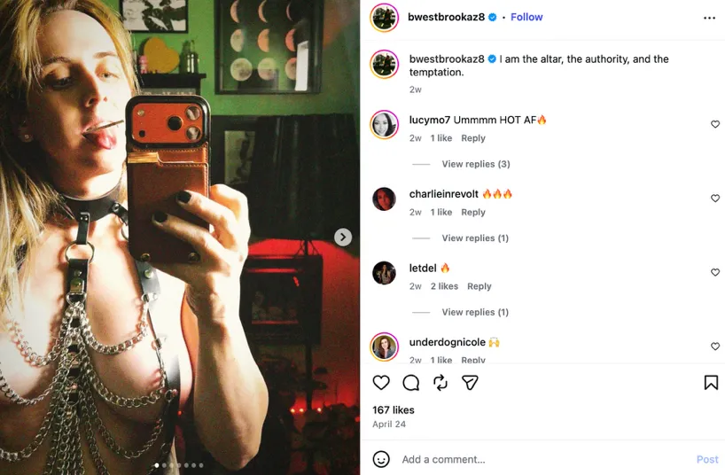 A person in a leather harness and chains takes a mirror selfie with their phone, displaying a lollipop in their mouth.