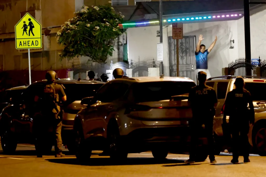 Police officers at night arresting a man with his hands raised on the porch of a building with glowing colorful lights.