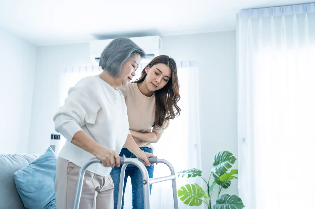 A younger woman helps an older woman use a walker at home.