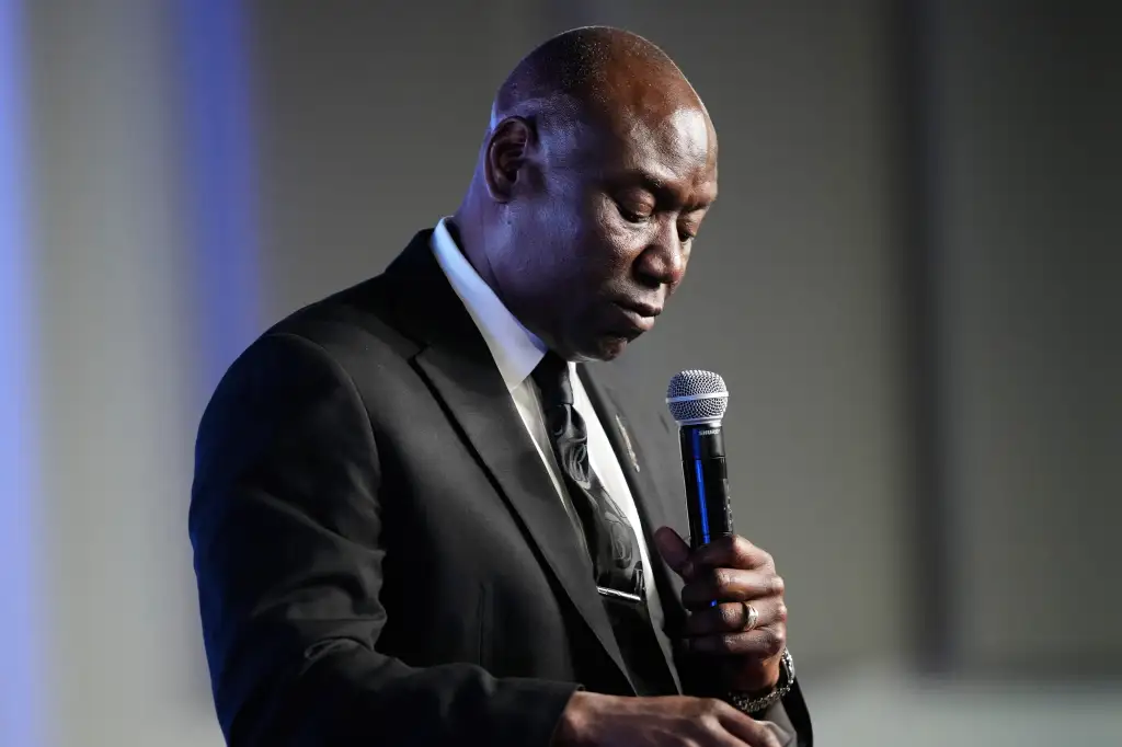 Ben Crump speaks at a memorial service, holding a microphone with his head bowed.